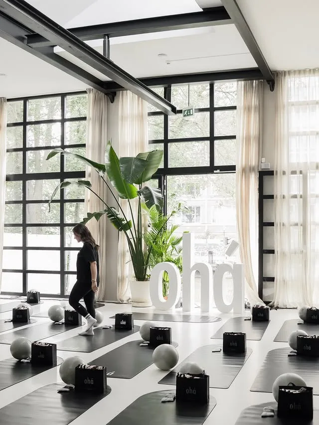 Şüheda walking through the ohá event room — forty black mats, goodie bags and the white ohá letters in daylight at Atelier Oost, Amsterdam